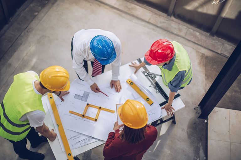 A group of contractors wearing protective gear gather around a table to discuss construction plans 